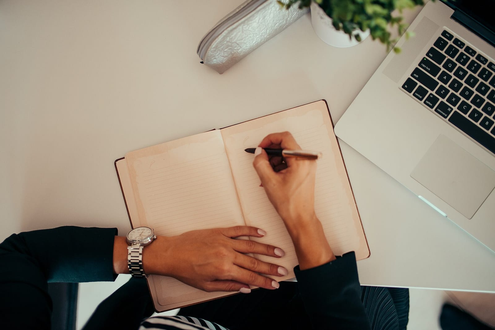يدان تكتبان في دفتر مفتوح على مكتب، وبجانبه حاسب محمول.